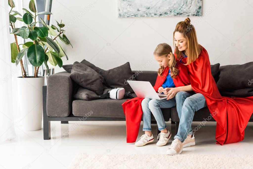 mother and daughter in red cloaks sitting on sofa and holding laptop