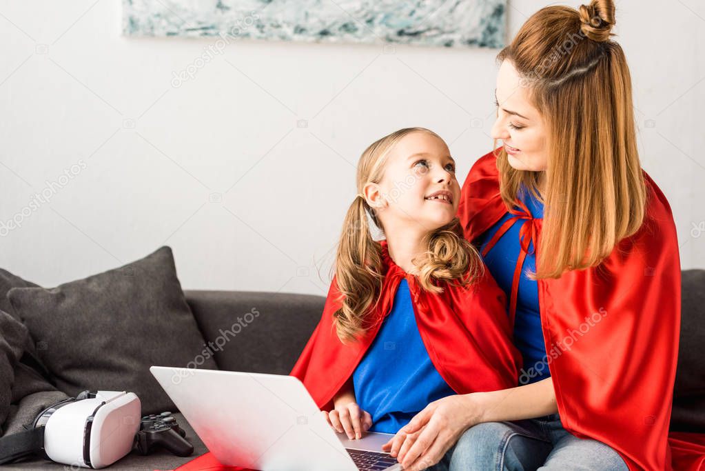 Cute kid and beautiful mother in red cloaks holding laptop and looking at each other