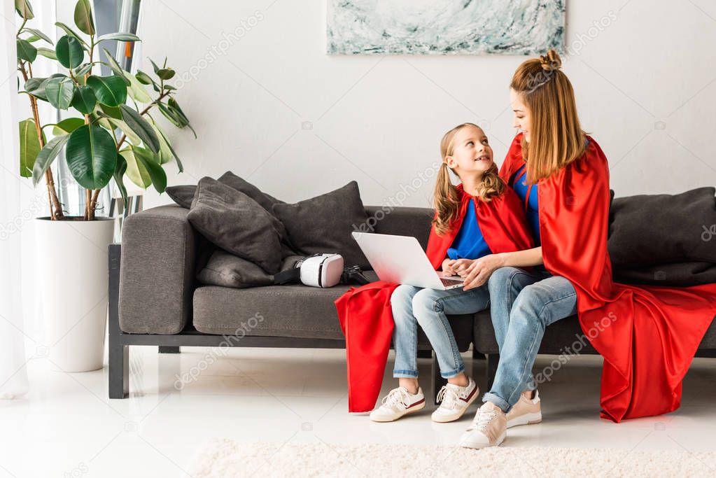 Cute kid and beautiful mother in red cloaks sitting on sofa and holding laptop