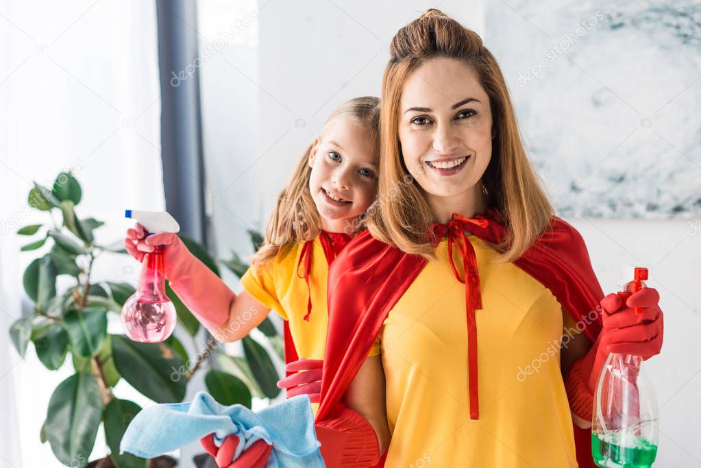 Mother and daughter in red capes and rubber gloves with sprays cleaning and looking at camera
