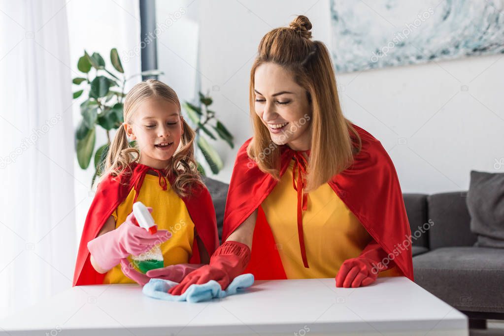 Mother and kid in red capes and rubber gloves dusting at home