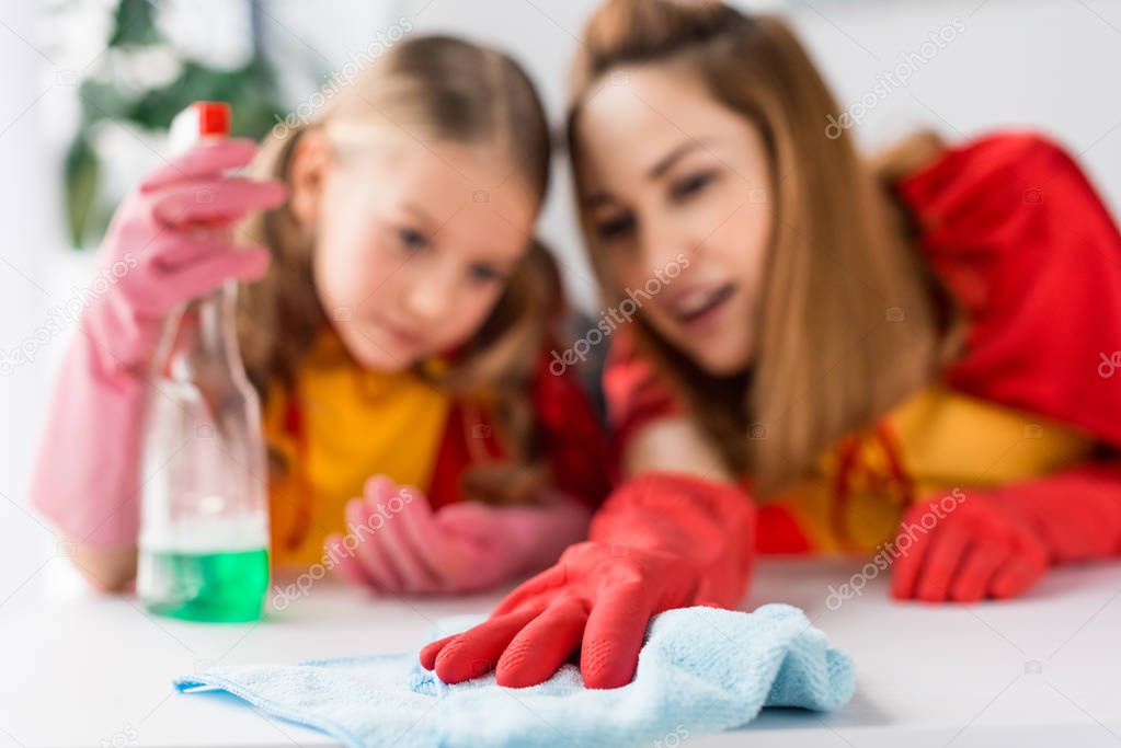 Selective focus of mother and daughter in red capes and rubber gloves dusting at home