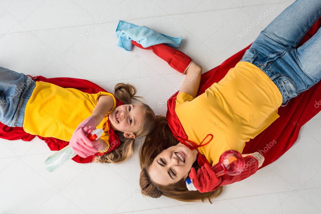 Top view of mother and kid in red capes and rubber gloves holding sprays and rug