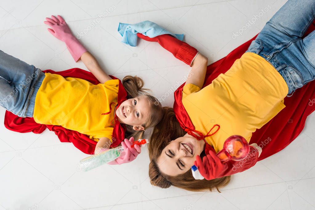Top view of smiling mother and kid in red capes and rubber gloves with sprays at home