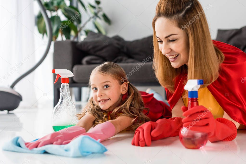 mother and cute kid in red capes and rubber gloves washing floor at home
