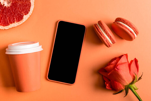 smartphone in coral case, coral paper cups, rose flower, macarons and grapefruit half on coral background, color of 2019 concept