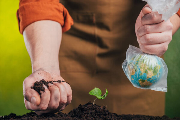 selective focus of man holding globe model in plastic bag and plating young green plant on blurred background, earth day concept