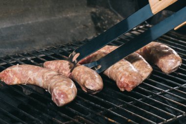 close up of tweezers near uncooked fresh sausages on bbq grill grates clipart