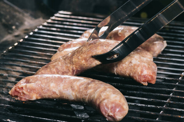 close up of tweezers near uncooked fresh sausages on bbq grill grates