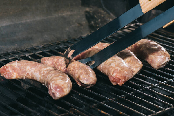 close up of tweezers near uncooked fresh sausages on bbq grill grates