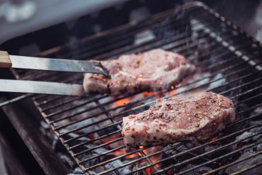 selective focus of juicy raw steaks grilling on barbecue grid with smoke and tweezers clipart