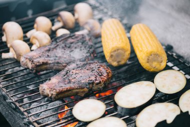 selective focus of juicy tasty steaks grilling on bbq grid with mushrooms, corn and sliced eggplant clipart