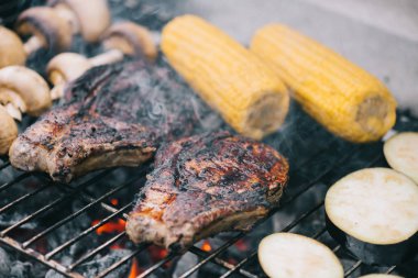 selective focus of juicy tasty steaks grilling on bbq grid with mushrooms, corn and sliced eggplant clipart