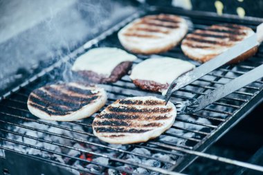 selective focus of tweezers and delicious fresh burgers ingredients with crust grilling on barbecue grid clipart