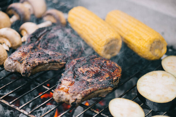 selective focus of juicy tasty steaks grilling on bbq grid with mushrooms, corn and sliced eggplant