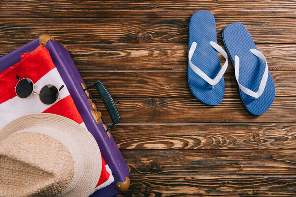 top view of summer accessories and flip flops near suitcase on wooden background