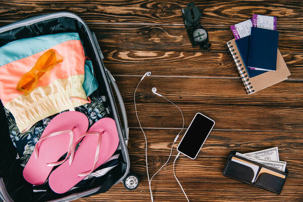 top view of smartphone with earphones, compass, passports with tickets and summer clothes in travel bag on wooden surface