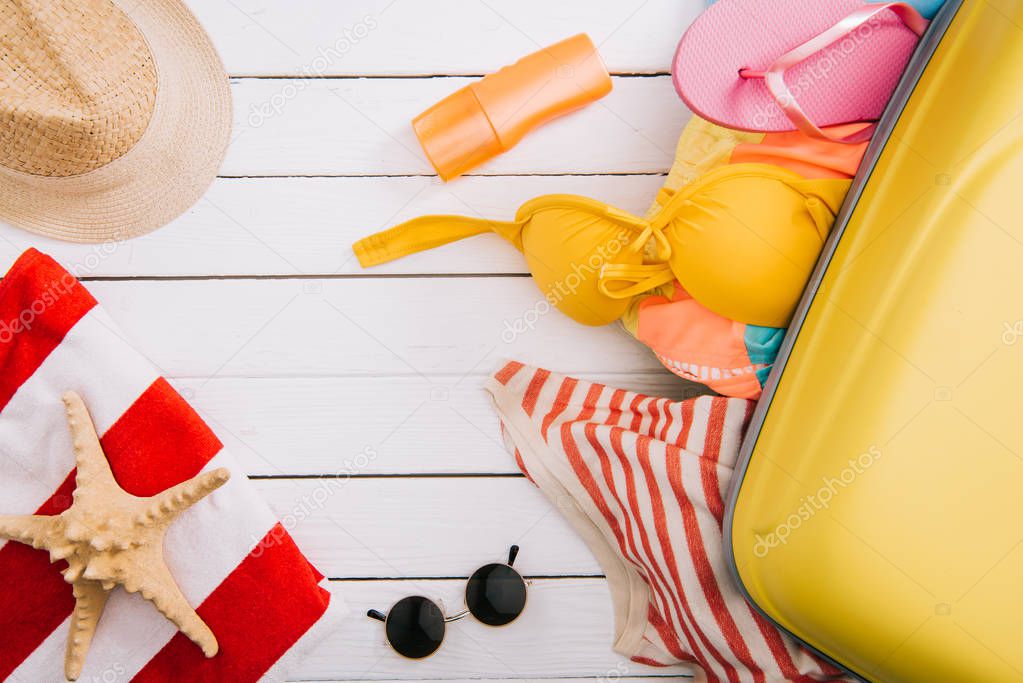 top view of sunscreen, starfish and summertime accessories and clothes in suitcase on white wooden background 