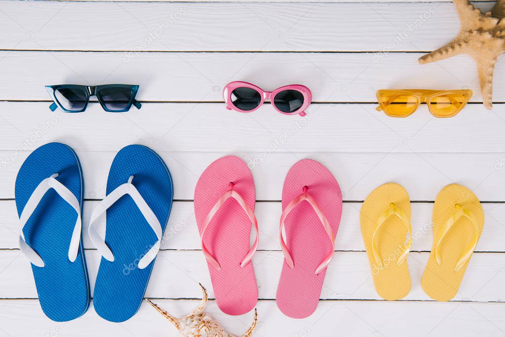 set of yellow, blue and pink sunglasses and flip flops on white wooden background 