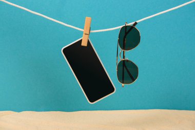 smartphone with blank screen and sunglasses hanging on rope with pin on blue background clipart