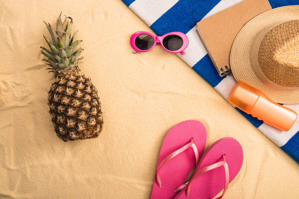 top view of summer accessories, sunscreen on striped towel and pineapple on sand