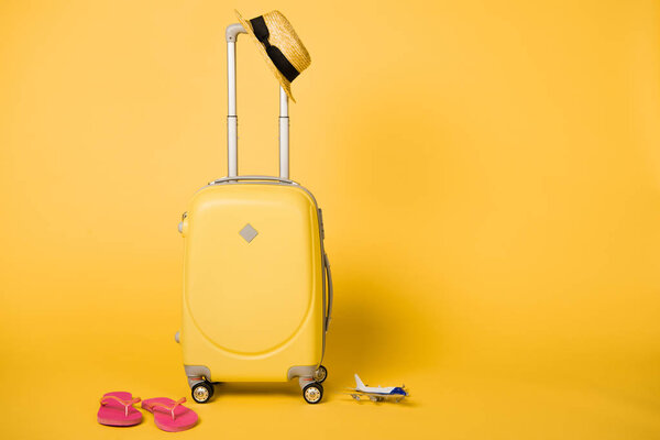 bright yellow travel bag, straw hat, pink flip flops and plane model on yellow background
