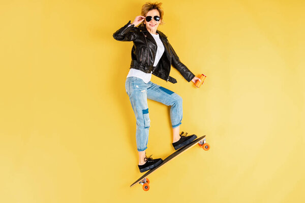 Confident girl standing on longboard and touching sunglasses on yellow background