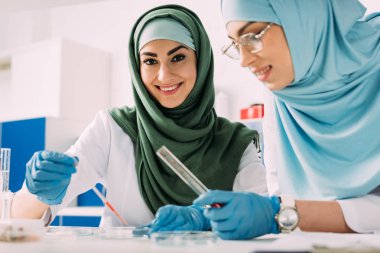 smiling female muslim scientists holding pipette and glass test tube during experiment in chemical lab clipart