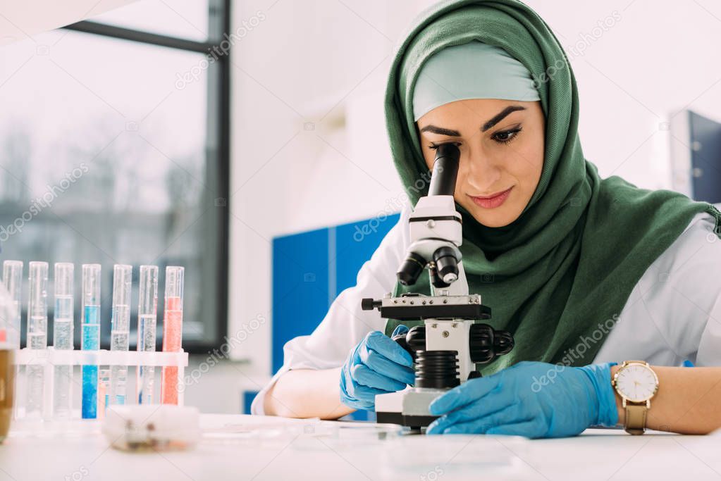 beautiful female muslim scientist looking through microscope during experiment in chemical laboratory