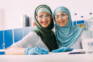koruyucu gözlük ve laboratuvar kamerada bakarak hijab gülümseyen kadın Müslüman kimyagerler