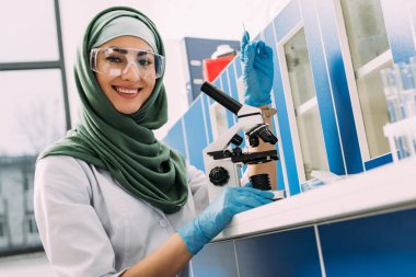 mikroskopla masada oturan, cam örnek tutarak ve kimyasal laboratuvar kamerada bakarak kadın Müslüman bilim adamı