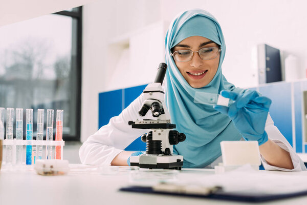 smiling female muslim scientist sitting at table with microscope and looking at glass sample during experiment in chemical laboratory