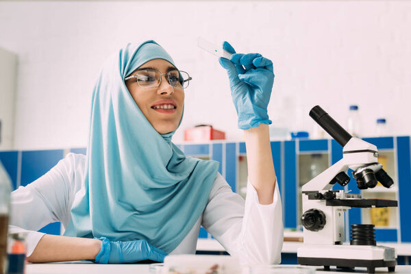 smiling female muslim scientist sitting at table with microscope and looking at glass sample during experiment in chemical laboratory