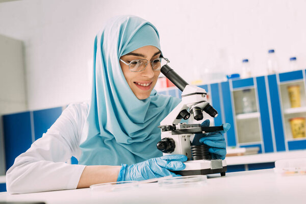 smiling female muslim scientist looking through microscope during experiment in chemical laboratory