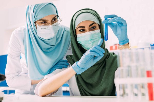 female muslim scientists in medical masks pointing with finger at glass test tube with reagent during experiment in chemical lab