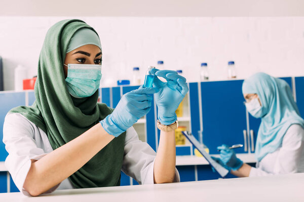мусульманка-ученый в медицинской маске держит пробирку с жидкостью во время эксперимента в химической лаборатории

