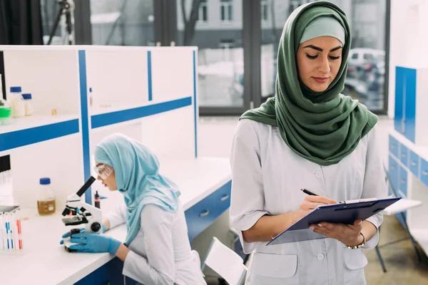 Mulheres Cientistas Muçulmanas Concentradas Usando Microscópio Escrita Prancheta Durante Experimento — Fotografia de Stock