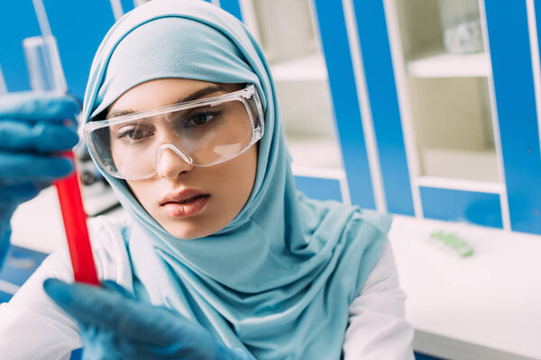 concentrated female muslim scientist in goggles holding test tube with red liquid in laboratory
