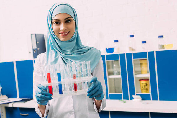 smiling female muslim scientist holding test tubes with red and blue liquid while looking at camera