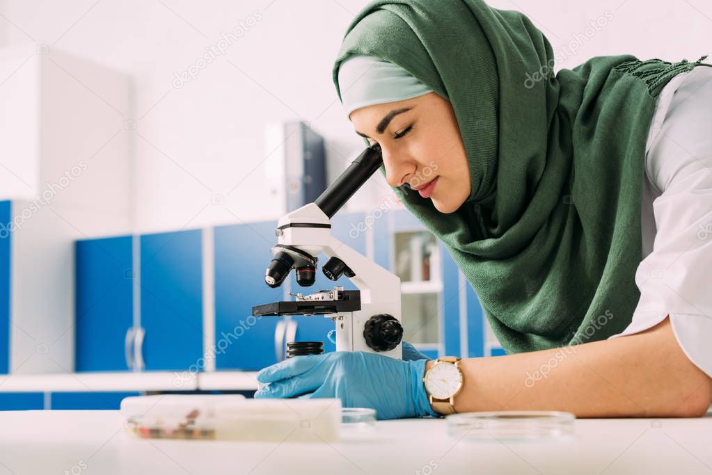 concentrated female muslim scientist looking through microscope during experiment in chemical laboratory