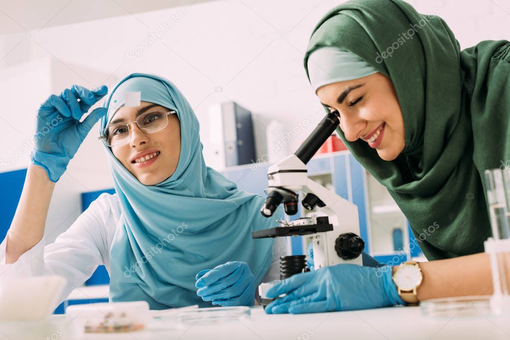 smiling female muslim scientists with microscope and glass sample during experiment in chemical laboratory