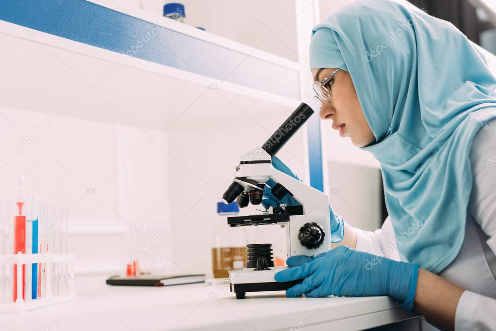 female muslim scientist looking through microscope during experiment in chemical laboratory with copy space