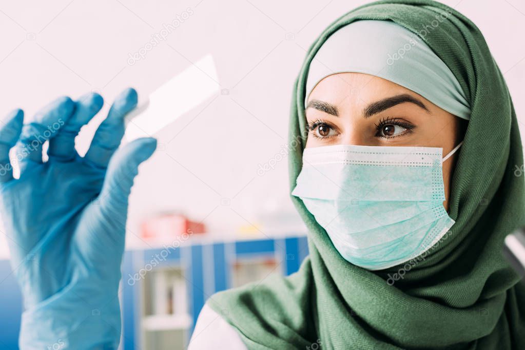 female muslim scientist in medical mask and hijab holding glass sample during experiment in laboratory