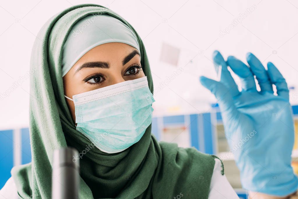 female muslim scientist in medical mask and hijab holding glass sample during experiment in laboratory