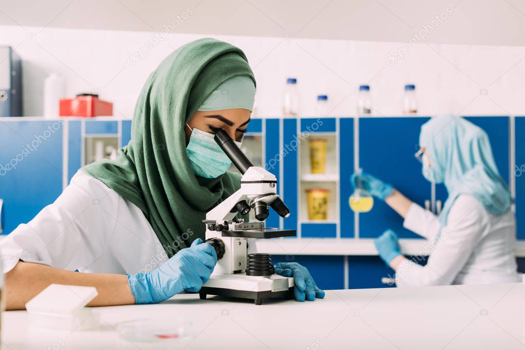 female muslim scientist looking through microscope during experiment with colleague working on background in chemical laboratory