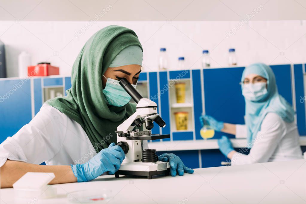 female muslim scientist looking through microscope during experiment with colleague working on background in chemical laboratory