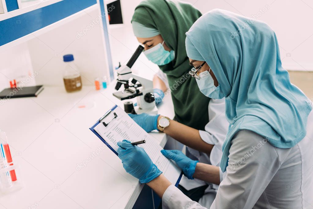 female muslim scientists in medical masks using microscope and clipboard during experiment in chemical laboratory