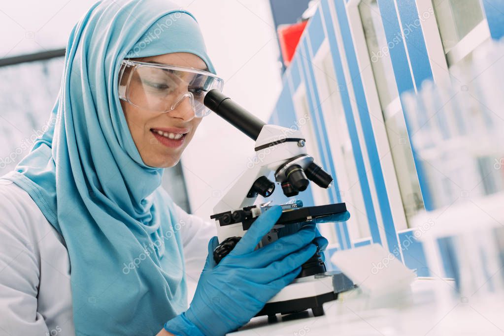 smiling female muslim scientist looking through microscope during experiment in chemical laboratory