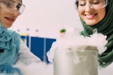 female muslim scientists in goggles experimenting with dry ice in chemical laboratory clipart