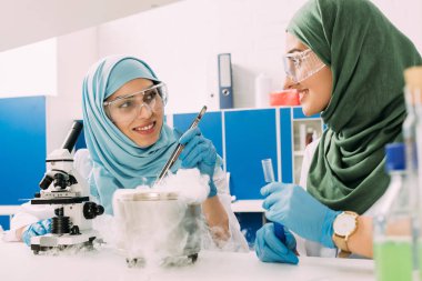 smiling female muslim scientists experimenting with microscope and dry ice in chemical laboratory clipart